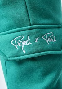 Grøn sweatshirtstof med en glat tekstur, der har hvid broderet tekst "Project x Paris" på brystområdet.
