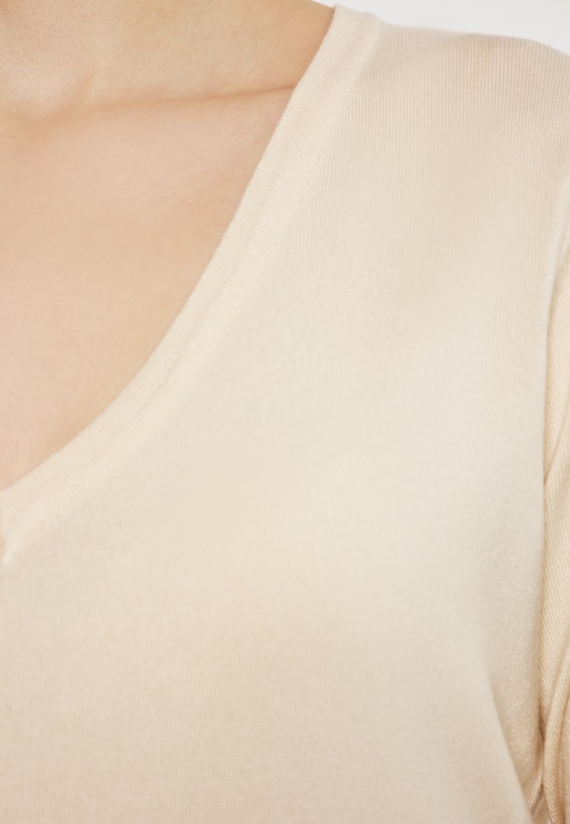 Beige V-hals top met ribbelstructuur, korte mouwen en een getailleerde pasvorm. Zachte stof valt soepel langs de halslijn en schouders.