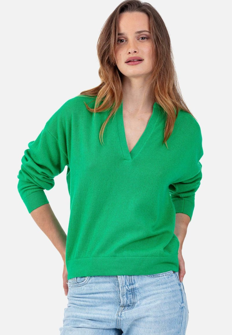 Grüner Strickpullover mit V-Ausschnitt und langen Ärmeln. Verfügt über eine lockere Passform sowie gerippte Bündchen und Saum. Kombiniert mit hellblauen Jeans.