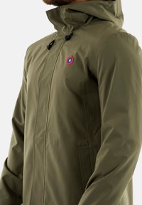 Veste imperméable vert olive avec une fermeture éclair à l'avant, une capuche ajustable et un logo rond rouge et bleu sur la poitrine. Texture lisse.