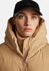 Doudoune beige avec un col montant, fermeture éclair, sous un col roulé beige côtelé. Casquette noire avec un logo brodé.