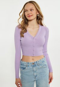 Cardigan a coste lilla con scollo a V, maniche lunghe e chiusura a cinque bottoni. Design cropped. Abbinato a jeans chiari a vita alta.