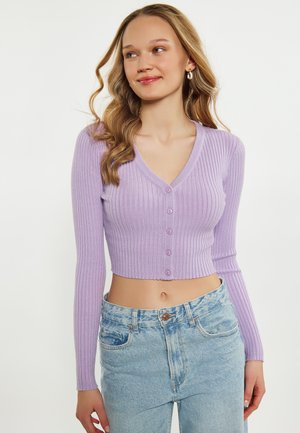 Cardigan a coste lilla con scollo a V, maniche lunghe e chiusura a cinque bottoni. Design cropped. Abbinato a jeans chiari a vita alta.