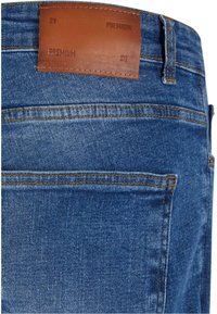 Jeans de mezclilla azul con una etiqueta de cuero en la cinturilla. Las características incluyen costuras naranjas y un diseño clásico de cinco bolsillos.