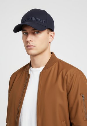 Jeune homme portant une casquette marine avec le logo "KARL LAGERFELD", une veste marron à fermeture éclair et une chemise blanche sur un fond clair uni.