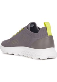 Zapatilla deportiva gris con parte superior de punto transpirable, suela de goma blanca y acentos amarillos. La parte trasera tiene un contrafuerte robusto para mayor soporte.
