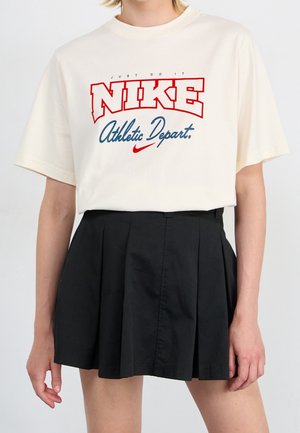 Personne portant un t-shirt blanc Nike avec un texte rouge et bleu et une jupe plissée noire, debout devant un fond uni.