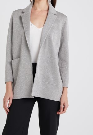 Femme portant un blazer argenté pailleté sur un haut blanc et un pantalon noir, avec de grandes boucles d'oreilles pendantes, sur un fond uni.