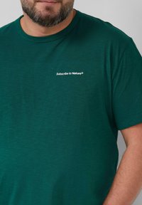 Dunkelgrünes T-Shirt mit kurzen Ärmeln aus weichem Stoff. Mit dem Aufdruck "Subscribe to Nature®" in Weiß auf der linken Brustseite.