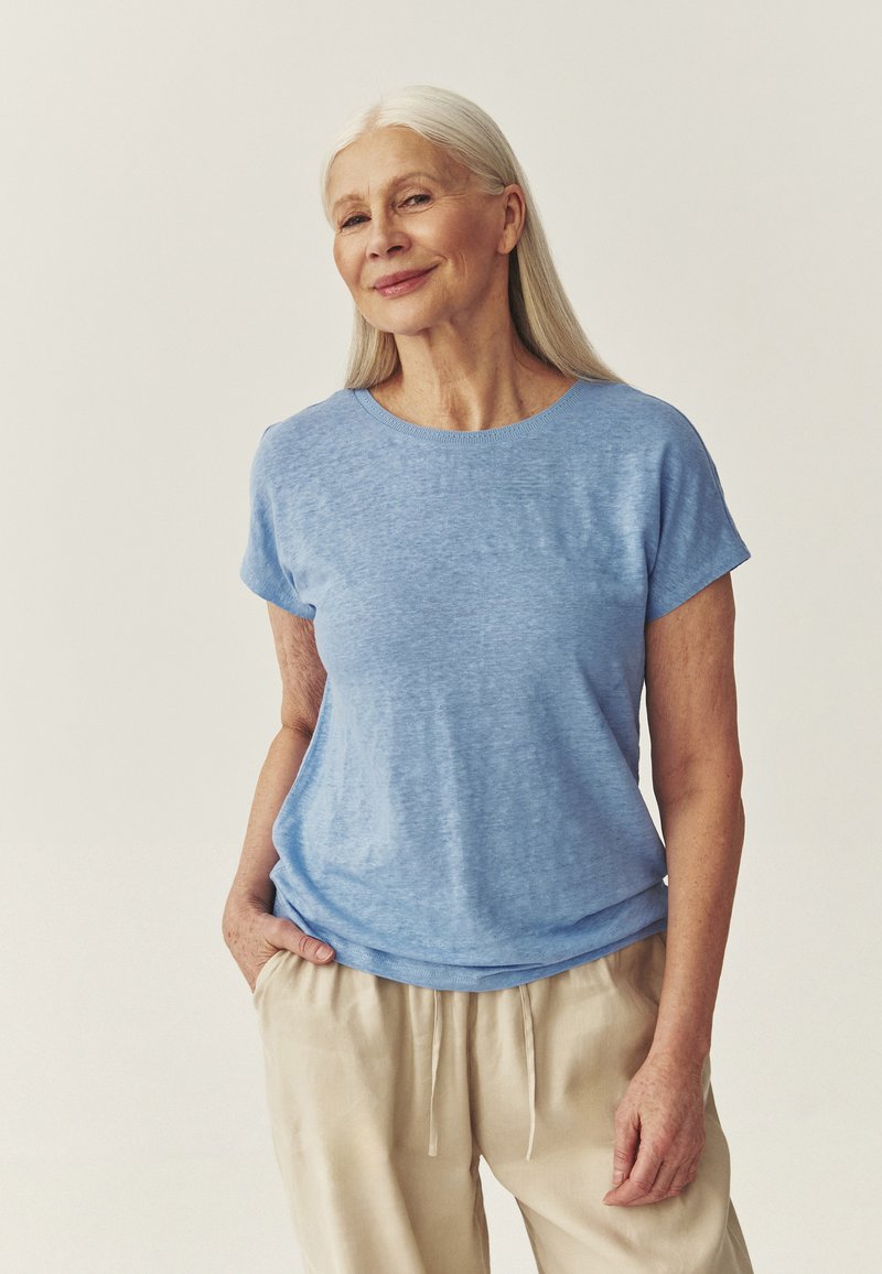 Femme âgée aux longs cheveux gris, portant une chemise bleu clair à manches courtes et un pantalon beige, debout les mains dans les poches, souriant doucement.