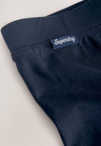 Nærbillede af mørkeblå stof med påsyet mærkat med teksten "Superdry" i hvide kursivbogstaver nær sømmen.