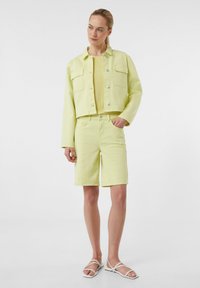 Jasnożółta dżinsowa kurtka z dwoma kieszeniami na piersi, w zestawieniu z pasującymi szortami. Pod spód założona teksturowana bluzka. Białe sandały na paski.