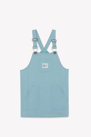 Light blue denim overall dress with cross-back straps, two front pockets, and metal hardware accents. Tag label positioned on the front.