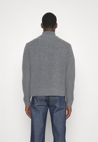 Man med ryggen vänd mot kameran, iförd en grå ribbad stickad tröja och blå jeans framför en enfärgad vit bakgrund.