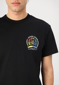 Svart bomulls T-skjorte med en fargerik brodert logo med sirkelmønster, tekst og laurbærakenter på venstre brystområde.