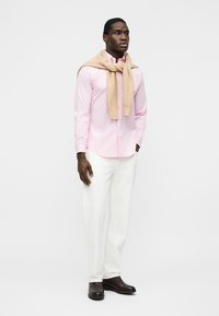 Muž v svetloružovej košeli na gombíky, béžovom svetri prehodenom cez plecia, bielych nohaviciach a tmavohnedých topánkach stojaci na jednoduchom pozadí.