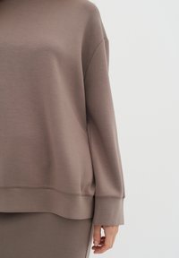 Sudadera marrón de corte holgado, textura suave y mangas largas. Presenta un cuello y dobladillo acanalados. Se muestra parcialmente cubriendo un conjunto a juego.