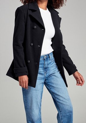 Femme portant un manteau noir croisé sur un top blanc et un jean bleu clair, debout devant un fond gris uni.