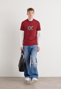 Piros pamut póló, multicolor "CK" logóval, fehér hosszú ujjú ing fölé rétegezve. Világoskék, kopott farmernadrággal és fekete táskával viselve.