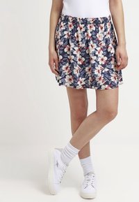 Woman wearing a navy skirt with white and red floral print, white socks, white sneakers, and a white top, standing with legs crossed.