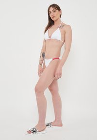Bikini bianco con top triangolare e slip a vita bassa, con accenti rossi. Il modello indossa ciabatte a stampa bianco e nero. Texture liscia.