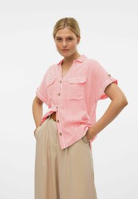Camisa de mangas cortas a rayas rojas y blancas con dos bolsillos frontales y botones dorados, confeccionada en tela ligera. Llevada con pantalones beige.