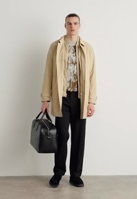 Beige trench coat egy textúrált szürke ing felett, fekete nadrág és fekete sportcipő mellett. Fekete bőr táskát tart, fémes díszítésekkel.