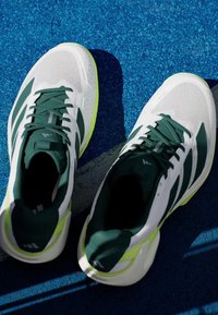 Bijele sportske tenisice s zelenim detaljima i vezicama, koje imaju prozračnu mrežastu gornjišnu konstrukciju, teksturirani đon i živopisni zeleni detalj na peti.