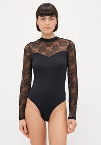 Femeie cu păr scurt și închis la culoare, purtând un body negru din dantelă cu mâneci lungi și un design floral transparent pe mâneci și partea superioară a pieptului.