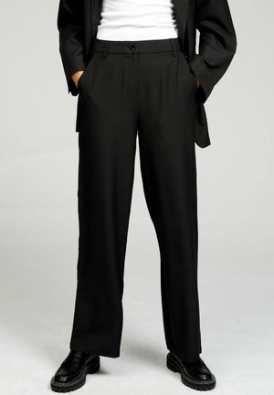 Person iført sorte skræddersyede bukser, sort blazer, hvid top og sorte loafers, hænder i lommerne, stående på lys grå baggrund.