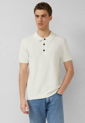 Jonge man draagt een witte, kortärmelig, gestructureerde polo met drie zwarte knopen en blauwe spijkerbroek, staand tegen een lichtgrijze achtergrond.