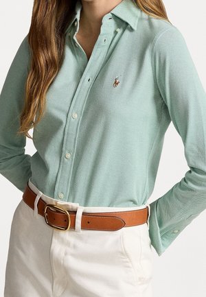 Femme portant une chemise boutonnée vert clair avec une ceinture marron et un pantalon blanc, les mains dans les poches, sur un fond uni.