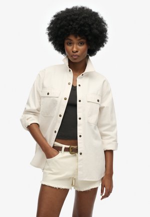 Lys beige jakke med to forlommer, knaplukning og rullede ærmer, parret med en mørk top og off-white shorts.