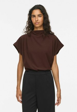 Femme aux longs cheveux bouclés portant un haut marron foncé sans manches et un pantalon noir taille haute, debout devant un fond blanc uni.