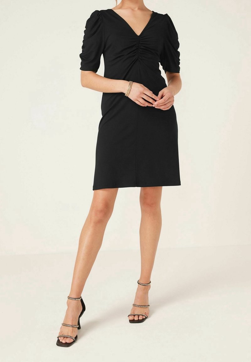 Robe noire avec un décolleté en V et un détail froncé au niveau de la poitrine, dotée de manches bouffantes et d'une silhouette ajustée, associée à des talons à lanières.