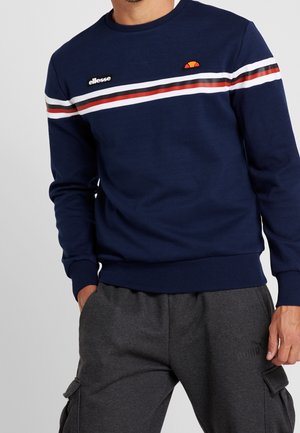 Mężczyzna ubrany w granatową bluzę z białymi, czerwonymi i czarnymi paskami na klatce piersiowej oraz szare spodnie cargo z kieszeniami.