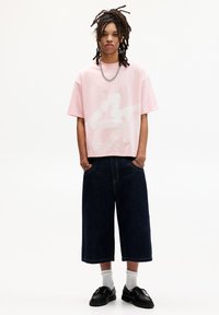 Vaaleanpunainen oversized t-paita, jossa on valkoinen grafiikka, yhdistetty tummiin denim-culotteihin. Mustat kengät ja valkoiset sukat viimeistelevät asun.