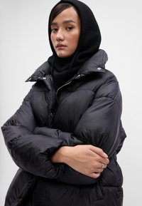 Casaco puffer preto com um colarinho alto e ombros grandes e acolchoados. Usado sobre um cachecol preto de malha grossa. Textura lisa e brilhante.