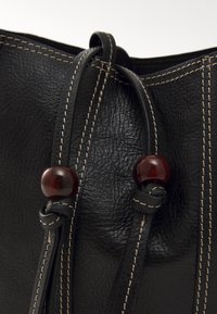 Primo piano di una borsa in pelle nera con texture a grana visibile, cuciture in contrasto e palline sferiche di un rosso scuro sui lacci dei chiusure.