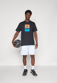 T-shirt noir à manches courtes avec un graphique de basket-ball coloré, associé à un short blanc et des chaussures de sport noires. Tenant un ballon de basket.