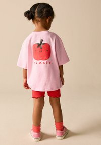 Růžové tričko s červeným grafickým motivem rajčete na zádech, v kombinaci s červenými žebrovanými šortkami. Oblečení je vyrobeno z měkké látky a má volný střih.