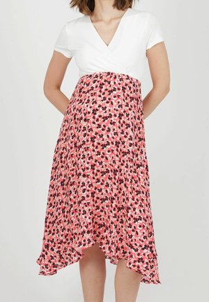 Woman wearing a white short-sleeve V-neck top and a pink floral high-low skirt against a plain background.