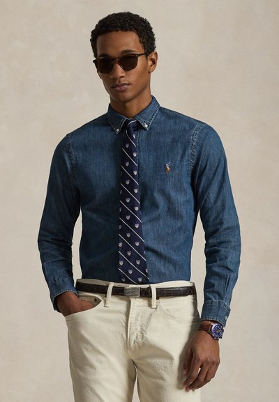 Uomo che indossa occhiali da sole scuri, una camicia di jeans blu abbottonata con una cravatta a righe, pantaloni color crema e un orologio con quadrante blu.