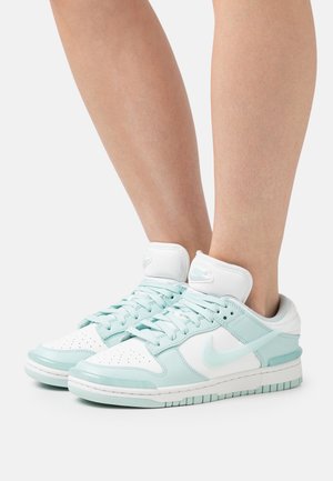 Stopy w białych i jasnoniebieskich sneakersach Nike z sznurówkami, widoczne kostki, na zwykłym białym tle.