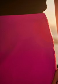 Fuchsiová sukně vyrobená z hladkého materiálu, s těsným střihem a černým pasem, s čistými okraji, částečně osvětlená slunečním světlem.