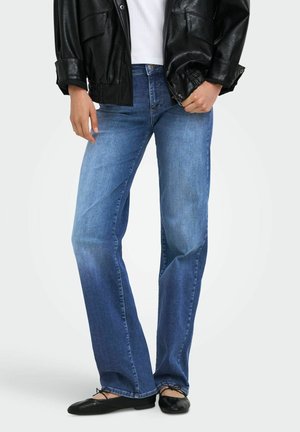 Person iført blå jeans med lige ben, sort læderjakke, hvid skjorte og sorte balletsko, der står mod en ensfarvet baggrund.
