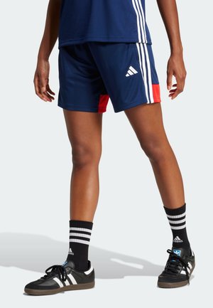 Persona que lleva pantalones cortos Adidas azul marino con rayas blancas y zapatos Adidas negros con calcetines a rayas negras y blancas.