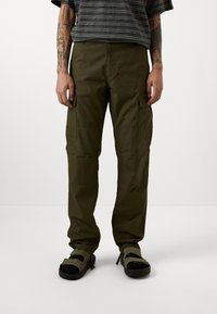 Oliwkowe spodnie bojówki z wieloma bocznymi kieszeniami, wykonane z trwałego materiału. Połączone z czarnymi sandałami z paskami na rzepy. Widać pasiastą koszulę.