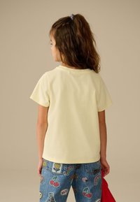 Dítě s dlouhými vlasy svázanými do culíku, oblečené v bledě žlutém tričku a modrých džínách ozdobených barevnými záplatami, stojící zády k pozorovateli na jednobarevném pozadí.
