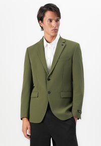 Young man with dark hair wearing an olive green blazer over a white shirt and black pants, standing with one hand in pocket.
